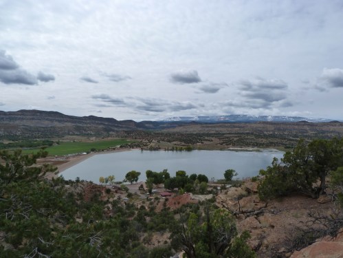 A scenic view of a lake surrounded by green fields and mountains under a cloudy sky.
