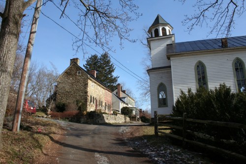 A scenic view of a rural road lined with historic stone and wooden buildings, including a church, under a clear blue sky.