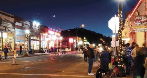 A bustling street at night, lined with colorful lights and shops, with people walking and a crescent moon in the sky.