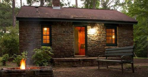 A cozy stone cabin with warm lights, surrounded by trees, and a fire pit in front.