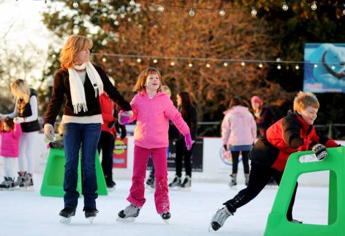 A woman and a girl skate on ice, while a boy practices with a skating aid. Others skate in the background.