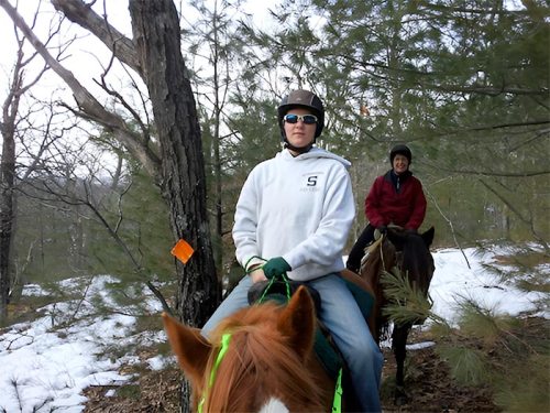 Two people riding horses through a snowy forest, one in a hoodie and sunglasses, the other in a red jacket.
