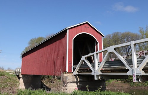 A red covered bridge with a metal support structure spans a small river, surrounded by greenery and blue sky.