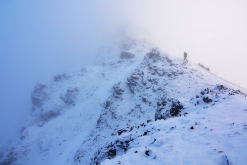 A snowy mountain peak shrouded in fog, with a lone hiker ascending the rocky terrain.