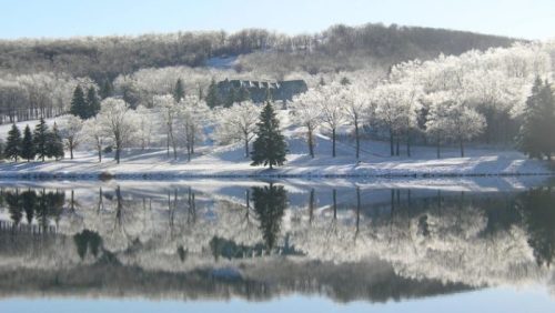 A serene winter landscape with frosted trees and a calm lake reflecting a snowy hillside.