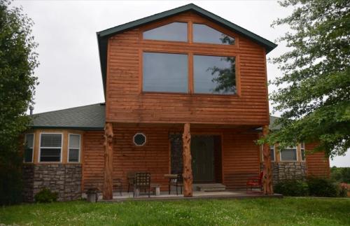 A two-story wooden house with large windows, stone accents, and a porch surrounded by greenery.