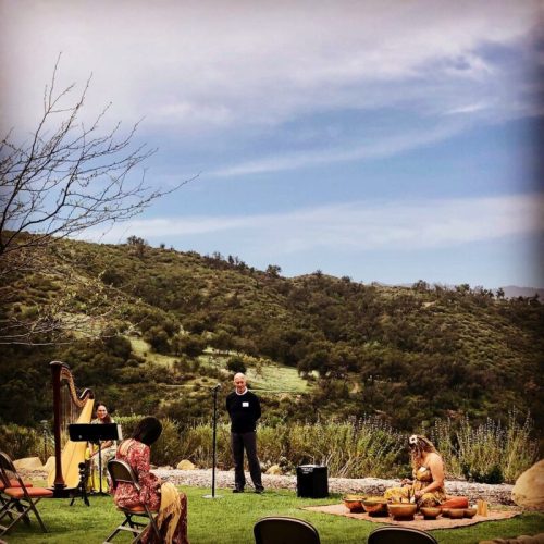 A serene outdoor gathering with a harpist, speaker, and participants seated on chairs, surrounded by lush hills.