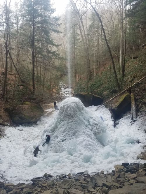 A frozen waterfall surrounded by trees, with people exploring the icy landscape below.