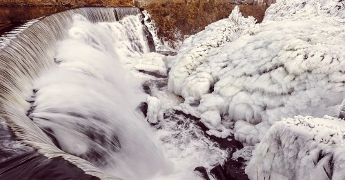 A frozen waterfall cascading over rocks, surrounded by ice and snow, creating a serene winter landscape.