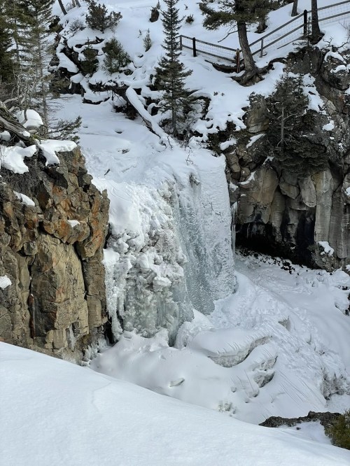 A frozen waterfall surrounded by snow-covered rocks and trees in a winter landscape.