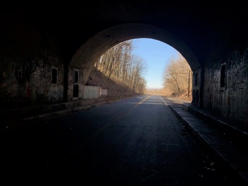A dark tunnel opening reveals a sunlit road lined with trees in the distance. Graffiti decorates the tunnel walls.