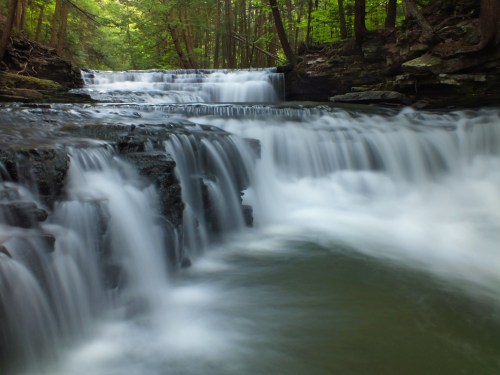 A serene waterfall cascades over rocky ledges, surrounded by lush green trees in a tranquil forest setting.