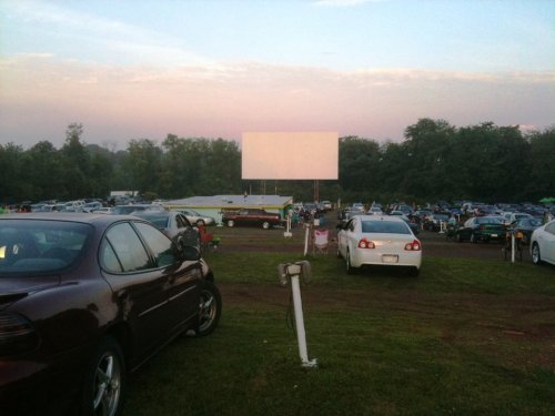 A drive-in movie theater with parked cars and a blank screen at dusk, surrounded by trees.