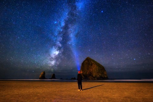 A person stands on a beach at night, gazing up at a starry sky with the Milky Way and a large rock formation in the background.