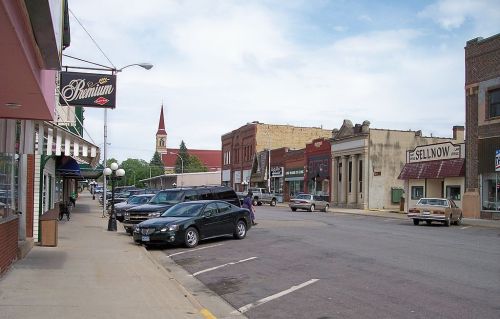 A small town street with shops, parked cars, and a church steeple in the background under a cloudy sky.