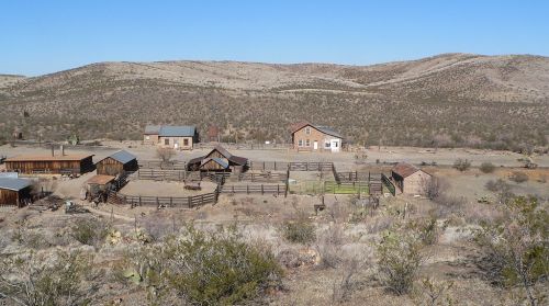 A panoramic view of a rustic ranch with several buildings and fenced areas, set against a hilly desert landscape.
