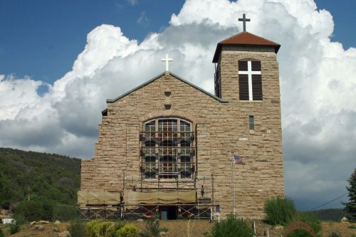 A stone church with a tall bell tower, under a cloudy sky, surrounded by scaffolding and greenery.