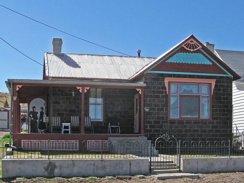 A historic stone house with a porch, decorative trim, and a fenced yard under a clear blue sky.