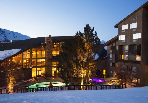 A cozy mountain resort at dusk, featuring a pool and hot tubs surrounded by snow-covered trees and buildings.