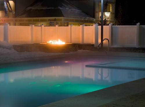 A serene night scene featuring a glowing pool, a fire pit, and a snowy landscape, with warm lights illuminating the area.