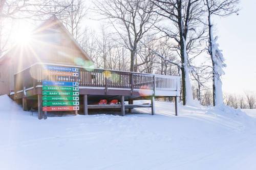 A ski lodge surrounded by snow-covered trees, with colorful trail signs and sunlight shining through.