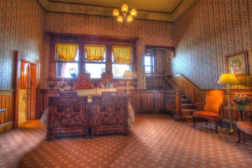 A vintage bedroom with ornate wooden furniture, patterned wallpaper, a staircase, and warm lighting.