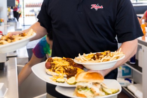 A server carries two plates of food, including fries and a sandwich, in a busy restaurant setting.