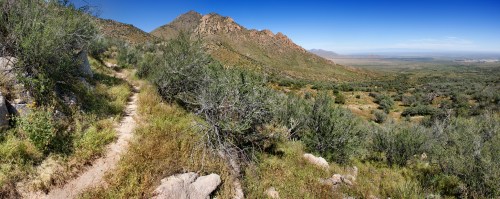 A panoramic view of a mountainous landscape with a winding trail, lush greenery, and a clear blue sky.