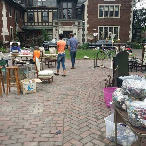 Two people walk through a yard sale with various items displayed on tables and the ground in a residential area.