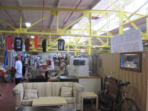 A spacious thrift store interior with colorful clothing, furniture, and a bicycle, featuring bright yellow beams overhead.