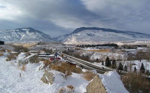 Snow-covered landscape with mountains in the background and a winding road in the foreground under a cloudy sky.