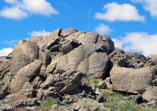 Ancient petroglyphs carved into large rocks, surrounded by grass and under a blue sky with fluffy clouds.