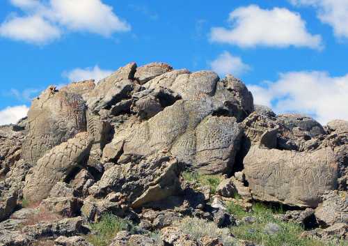 Ancient petroglyphs carved into large rocks, surrounded by sparse vegetation under a blue sky with clouds.