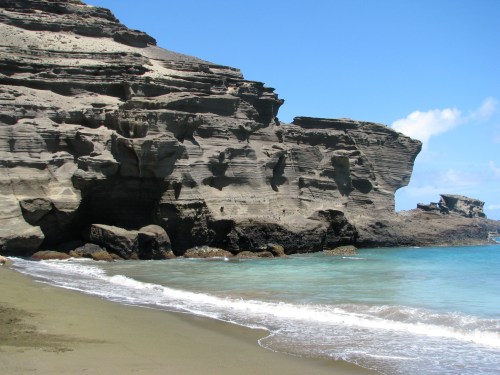 A rocky coastline with steep cliffs beside a calm turquoise sea and sandy beach under a clear blue sky.