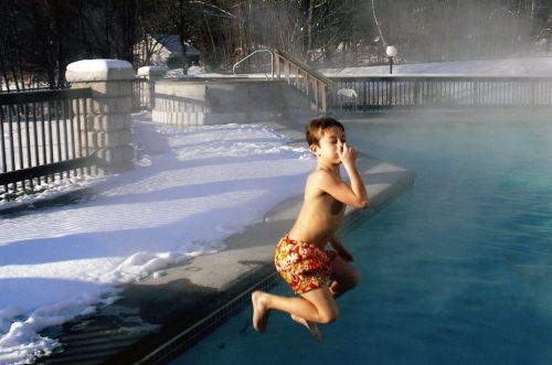 A boy in swim trunks jumps into a steaming pool surrounded by snow and a wooden fence.