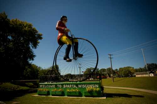 A large statue of a man on a penny-farthing bicycle welcomes visitors to Sparta, Wisconsin. Clear blue sky above.