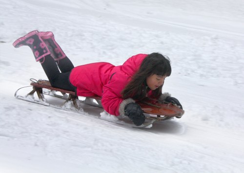 A girl in a pink coat and boots sleds down a snowy hill, lying on her stomach on a wooden sled.