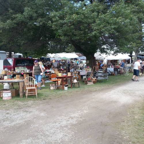 A bustling outdoor market with various stalls selling antiques and crafts, surrounded by trees and tents.