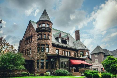 A large, historic stone mansion with a turret, surrounded by greenery and under a cloudy sky.