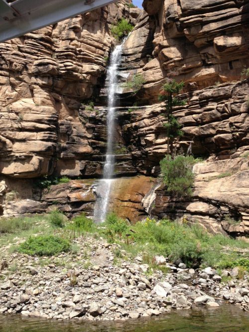 A waterfall cascading down rocky cliffs, surrounded by greenery and a rocky riverbank.