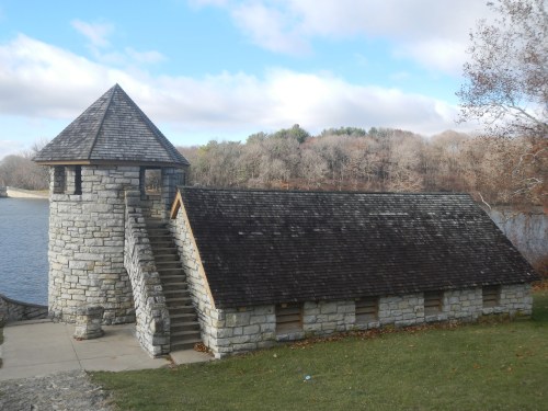 Stone building with a tower beside a lake, surrounded by trees and a cloudy sky. Steps lead up to the entrance.