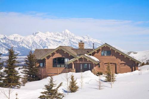 A cozy wooden cabin surrounded by snow, with mountains in the background under a clear blue sky.