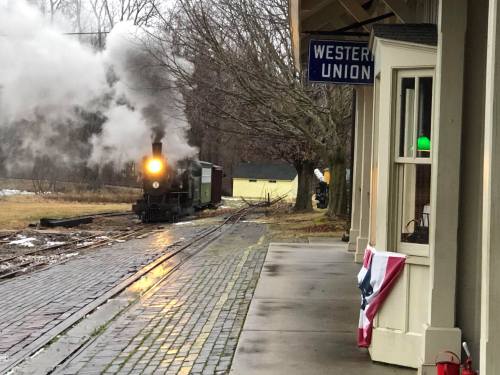 A steam train approaches a station with a Western Union sign, surrounded by trees and a flag draped nearby.