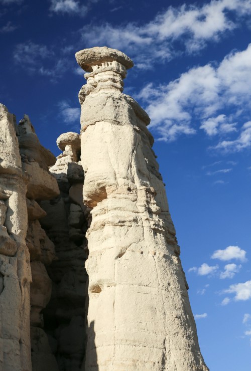 A tall, narrow rock formation rises against a blue sky with scattered clouds, showcasing unique geological features.