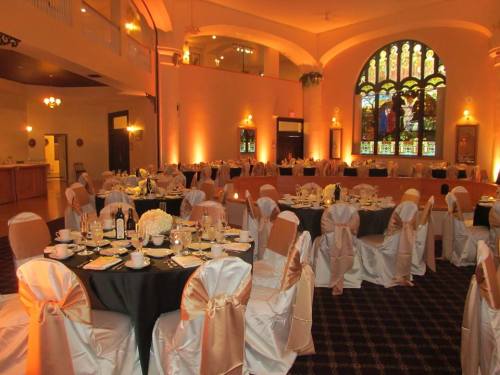 Elegant banquet hall set for a formal event, featuring round tables with white and gold decor and a stained glass window.
