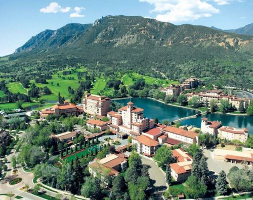 Aerial view of a resort surrounded by lush greenery, mountains, and a lake, with buildings and golf courses visible.