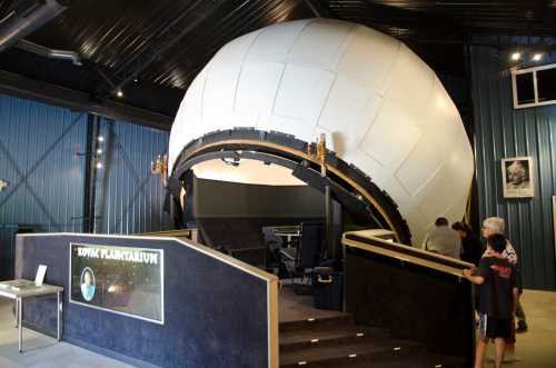 A large, white planetarium dome inside a building, with visitors approaching the entrance and a display area nearby.