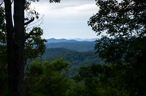 A scenic view of rolling hills and distant mountains framed by trees under a cloudy sky.