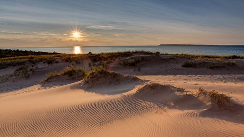 Sunset over a tranquil beach, with gentle sand dunes and grass in the foreground, reflecting warm golden hues.