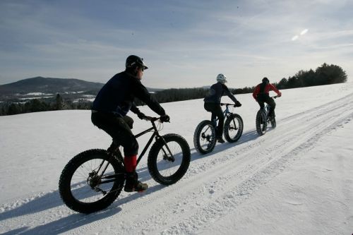 Three cyclists ride fat bikes on a snowy landscape, with mountains in the background under a clear blue sky.